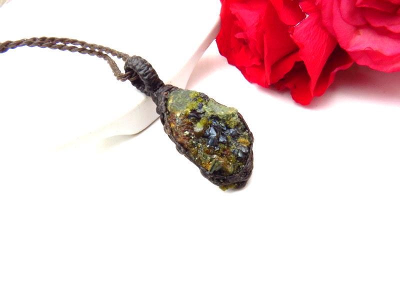 May include: A handcrafted necklace featuring a raw, green gemstone pendant wrapped in a dark brown, woven material. The pendant hangs from a matching brown cord. Pink and red roses are in the background, resting on a wooden surface.