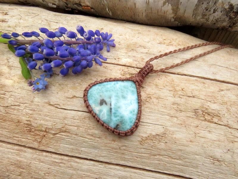 May include: A handmade necklace featuring a triangular, light blue stone pendant with brown macrame detailing. The pendant is suspended from a brown cord. A cluster of purple flowers rests on a wooden surface.