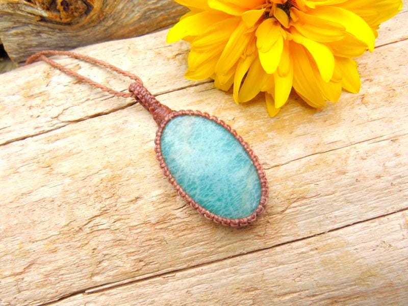 May include: A woman with long brown hair wearing a brown vest over a yellow tank top and a turquoise teardrop pendant necklace. She is standing in front of a body of water.