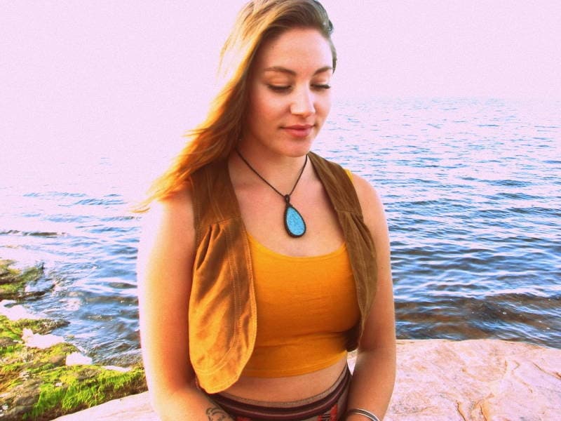May include: A woman with long brown hair wearing a brown vest over a yellow tank top and a turquoise teardrop pendant necklace. She is standing in front of a body of water.
